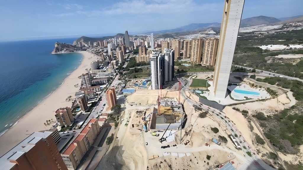 Aerial drone view of TM Tower construction site on Playa de Poniente Benidorm, April 2026, with foundation excavation, primary tower crane, INTEMPO at the right of frame and the Sunset towers along the beach