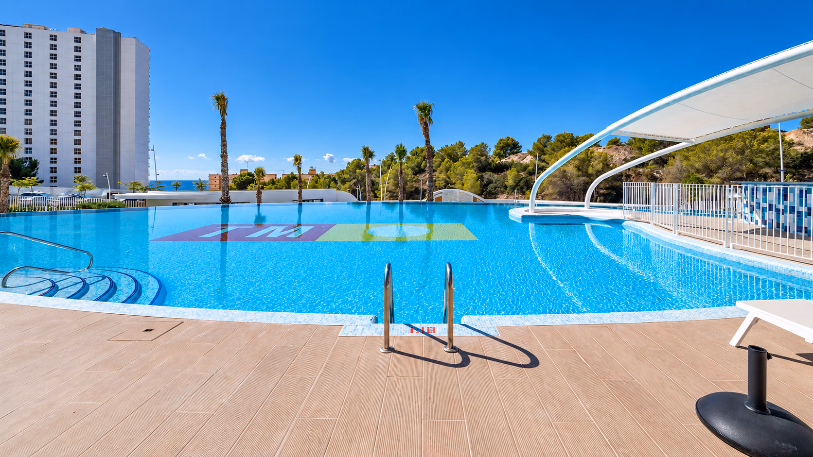 Large outdoor pool with a sun deck, palm trees and a shade canopy
